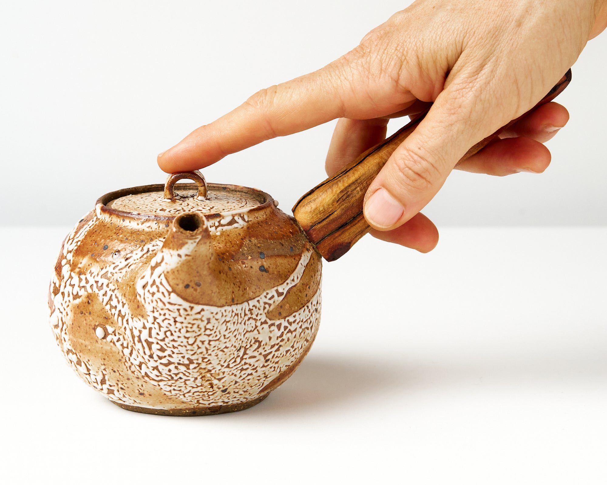 Ceramic teapot with wooden handle on a white background, with hand to show scale