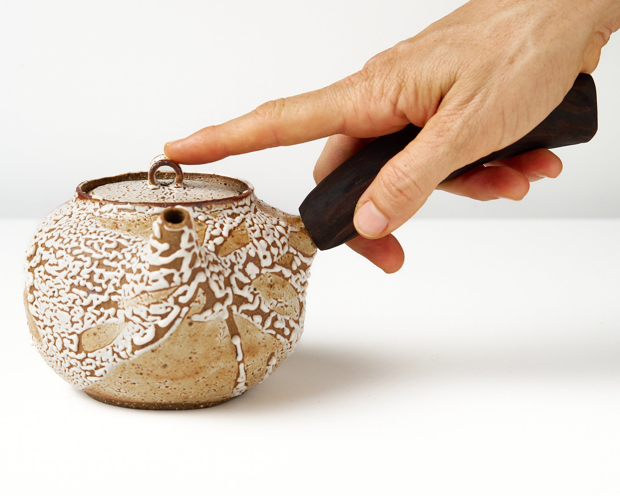 Ceramic teapot with a wooden handle on a white background, front view with hand to show scale